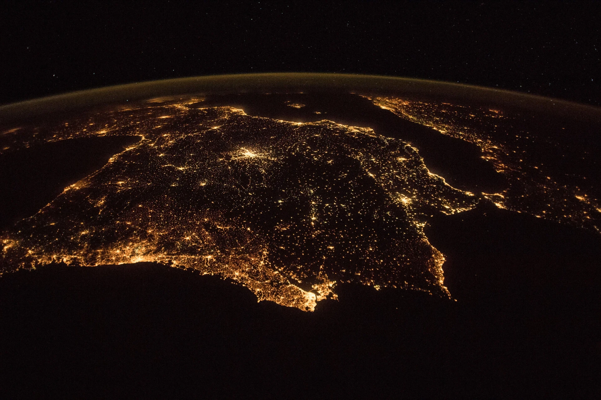 Night view of Earth from orbit, city lights tracing coastlines and inland grids in orange