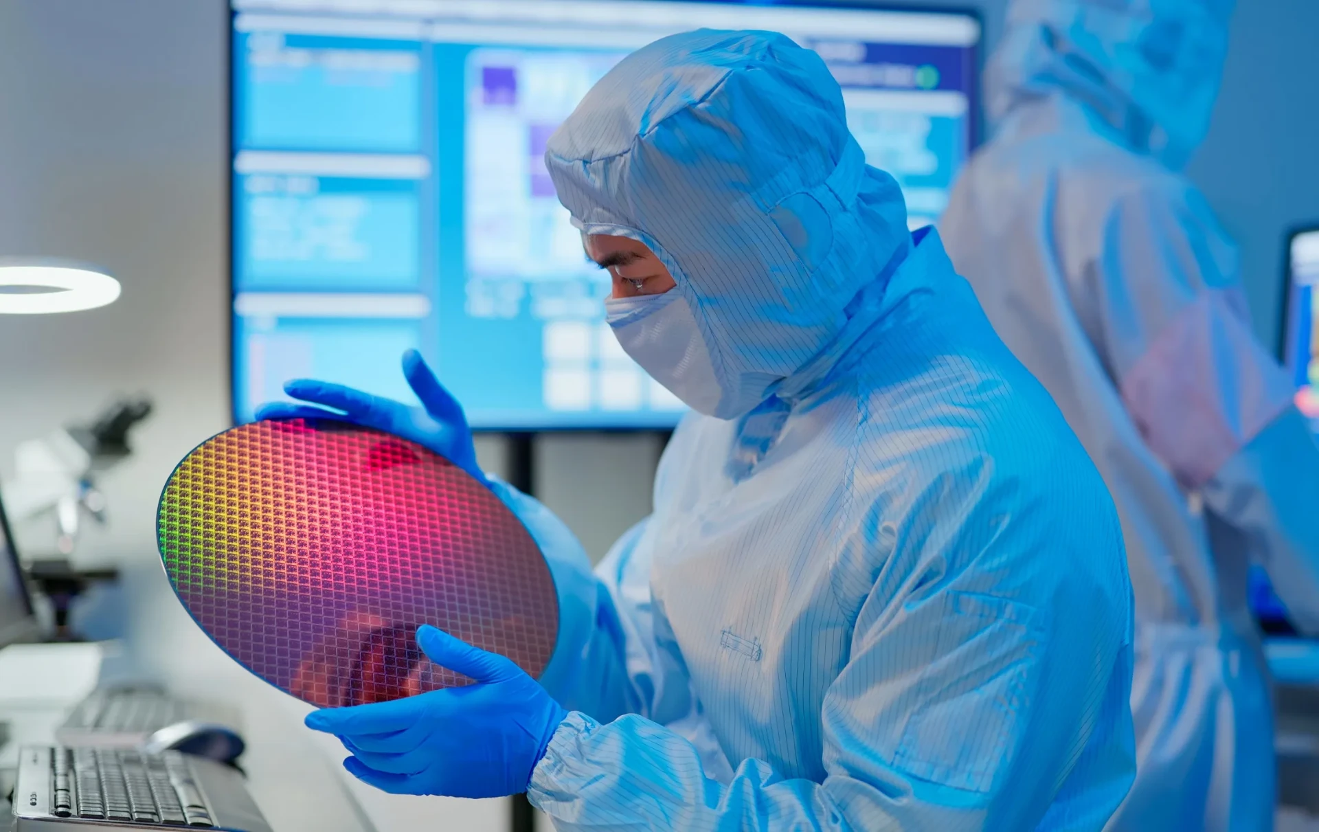 Engineer in a cleanroom suit inspecting a patterned silicon wafer beside process monitors